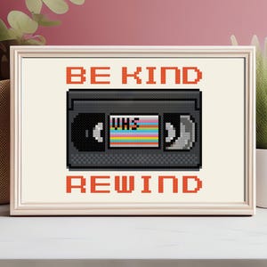 May include: A framed cross-stitch picture with a retro VHS tape design. The tape has a colourful label and is set against a cream background. The words "BE KIND REWIND" are stitched in orange above and below the tape.