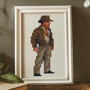 Op de afbeelding: Een ingelijste pixel art van een figuur met een bruine hoed, jas en broek. Het kunstwerk zit in een witte lijst en heeft een pixelachtig ontwerp, dat doet denken aan een klassiek videogamepersonage. De figuur staat tegen een gestippelde achtergrond.