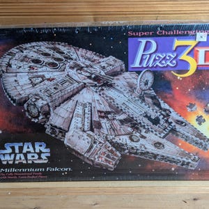 Puede incluir: Una caja de rompecabezas 3D de Star Wars Millennium Falcon. La caja es morada y negra con las palabras "Super Challenging" y "Puzz 3D" en blanco. La imagen en la caja muestra la nave espacial Millennium Falcon en el espacio.