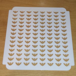 May include: White plastic stencil with a repeating pattern of bat silhouettes. The stencil is designed for crafting and decorating.