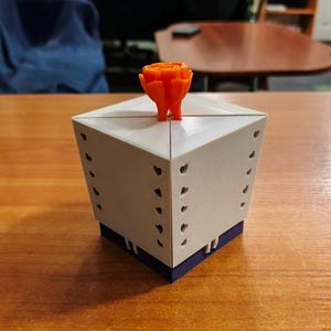 Könnte beinhalten: Eine geometrische, hellgraue Box mit herzförmigen Aussparungen und einem dunkelblauen Sockel. Ein orangefarbenes, rosenförmiges Objekt sitzt oben auf der Box. Die Box steht auf einer Holzoberfläche.