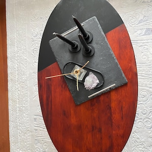 Puede incluir: Mesa de centro ovalada de madera con acabado negro y rojo. La mesa está decorada con una bandeja de pizarra negra con tres velas negras, una bandeja pequeña con un recipiente de vidrio y una flor decorativa.