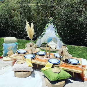 May include: Outdoor picnic setup with a low wooden table set for six. White plates with blue rims, yellow napkins, and decorative placemats are arranged. Pillows, cushions, and a small teepee add to the bohemian aesthetic. Pampas grass and candles complete the scene.