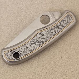May include: A folding pocket knife with a silver-toned metal handle featuring intricate black scrollwork engravings and a thin gold-colored border. The blade is partially visible, showing a circular thumb hole and the partial text Spyderco.
