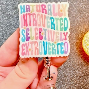 May include: A rectangular badge reel with the words "NATURALLY INTROVERTED SELECTIVELY EXTROVERTED" in colorful bubble letters. The badge has a white background with small flower accents and a retractable cord and clip.