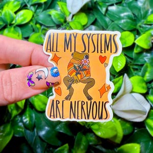 Puede incluir: Una pegatina con una rana de dibujos animados sosteniendo una flor y el texto "All my systems are nervous". La pegatina está sobre un fondo melocotón con corazones rosas, estrellas y un símbolo yin yang.