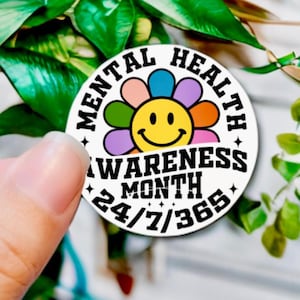 May include: White round sticker with a black border and the text "Mental Health Awareness Month 24/7/365". A colorful flower with a yellow smiley face is in the center of the sticker.