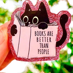 May include: A pink glittery cat-shaped pin with a black cat peeking over a pink book. The book reads "BOOKS ARE BETTER THAN PEOPLE" in black text. The pin has a pink glitter border and a black tail.