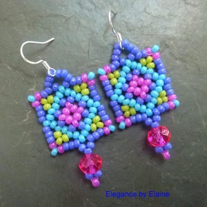 Op de afbeelding: Een paar kleurrijke kralen oorbellen met een geometrisch ontwerp. De oorbellen zijn gemaakt van blauwe, groene, roze en paarse kralen. Elke oorbel heeft een roze kristallen hanger.