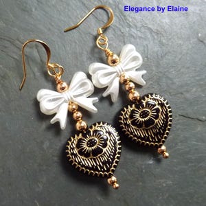 May include: Gold tone earrings with white pearl bow accents and black heart charms with gold detailing.  Elegance by Elaine.