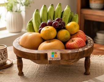 May include: A round wooden fruit bowl with four short legs and inlaid white floral patterns along the rim. The bowl is filled with a variety of fresh fruit including green bananas, red apples, oranges, and dark purple grapes. The text Craft Cooper is visible.