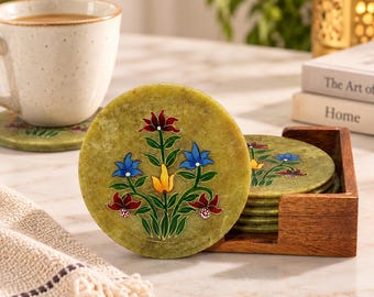 May include: A set of round, light green stone coasters featuring a hand-painted floral design with red, blue, and yellow petals and green stems. The coasters are stored in a square wooden holder, with one coaster displayed upright in front.