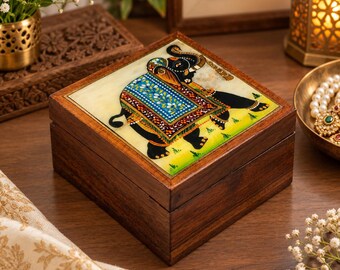 May include: A square wooden box featuring an inlaid stone panel on the lid. The panel displays a hand-painted black elephant adorned with a vibrant blue, red, and gold patterned blanket, set against a light yellow and cream-colored background.
