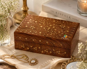 May include: A rectangular wooden box with a dark brown finish, featuring intricate gold-colored floral and vine inlay patterns on the lid and sides. The box has a smooth, polished surface and a small logo printed on the top right corner of the lid.