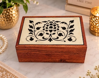 May include: A rectangular wooden box with a dark brown grain finish and a flat lid. The lid features an inset cream-colored panel decorated with a symmetrical black floral and vine pattern centered on the top surface of the closed storage container.