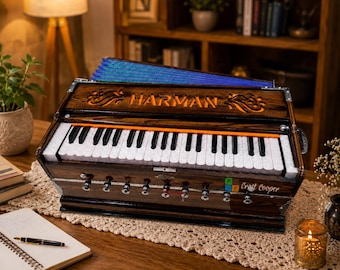 May include: A dark wood harmonium musical instrument featuring a keyboard with white and black keys, an orange accent strip, and a blue bellows section. The front panel displays the brand name HARMAN and the text Craft Cooper in white lettering.