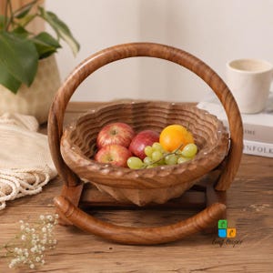 Op de afbeelding: Een ronde, inklapbare houten fruitmand met een geschulpte rand en een gebogen handvat, rustend op een houten voet. De mand bevat twee rode appels, een sinaasappel en een tros groene druiven. Het logo Craft Cooper is zichtbaar.