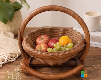 May include: A round, collapsible wooden fruit basket with a scalloped edge and a curved handle, resting on a wooden base. The basket contains two red apples, one orange, and a cluster of green grapes. The logo Craft Cooper is visible.