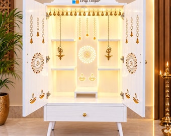 May include: A white wooden home temple cabinet with open doors, featuring gold-colored laser-cut Om symbols and diya lamp motifs. The interior includes shelves, hanging brass bells, and integrated warm lighting. The text Craft Cooper is visible.