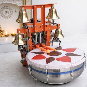 Puede incluir: Un instrumento musical naranja y plateado con cuatro campanas de latón y un tambor con una parte superior blanca decorada con formas de corazón rojas y marrones. Dos baquetas naranjas descansan sobre el tambor.