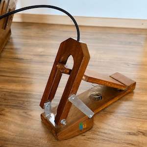 Puede incluir: Un pedal de pie de madera con acabado marrón, que cuenta con un mecanismo de resorte metálico, soportes de metal plateado y un cable negro que se extiende desde la parte superior. La base del pedal muestra un pequeño logo con el texto Craft Cooper en negro.