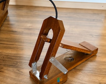 May include: A wooden foot pedal with a brown finish, featuring a metal spring mechanism, silver-colored metal brackets, and a black cable extending from the top. The base of the pedal displays a small logo with the text Craft Cooper in black.