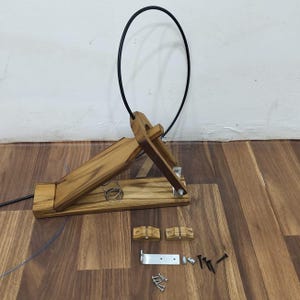 Puede incluir: Una honda de madera con una banda circular negra, herrajes metálicos y componentes adicionales de madera y metal. La honda está hecha de madera de color marrón claro y está parcialmente ensamblada. Los componentes están dispuestos sobre una superficie de madera.