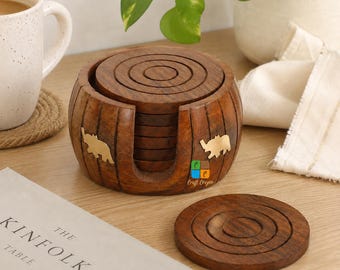May include: A set of round dark brown wooden coasters stored in a matching cylindrical wooden holder. The holder features brass elephant inlays on the exterior. One coaster is placed flat in front of the holder, displaying concentric circular grooves.