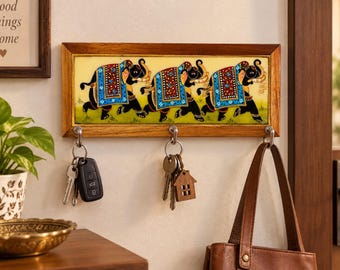 May include: A rectangular wooden wall-mounted key holder featuring a central tile painted with three black elephants adorned in colorful patterned blankets. The frame includes three silver metal hooks, each holding sets of keys and a brown bag.