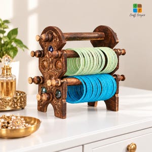 May include: A dark brown wooden bangle holder with intricate carved details and inlaid colorful glass accents. The stand features two horizontal rods holding a stack of light green bangles on the top rod and bright blue bangles on the bottom rod.