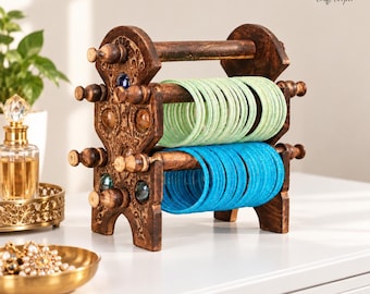 May include: A dark brown wooden bangle holder with intricate carved details and inlaid colorful glass accents. The stand features two horizontal rods holding a stack of light green bangles on the top rod and bright blue bangles on the bottom rod.