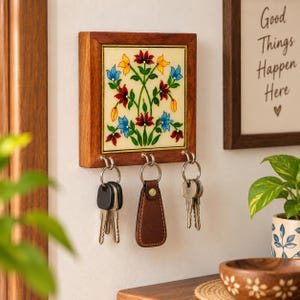 May include: A square wooden wall-mounted key holder featuring a central tile with a hand-painted floral design in red, blue, and yellow. The frame has three metal hooks, each holding a set of keys, including one with a brown leather fob.