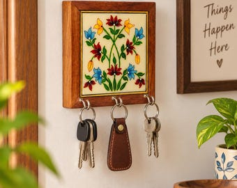 May include: A square wooden wall-mounted key holder featuring a central tile with a hand-painted floral design in red, blue, and yellow. The frame has three metal hooks, each holding a set of keys, including one with a brown leather fob.