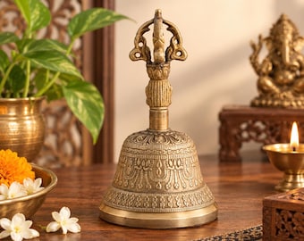 May include: A brass hand bell featuring an ornate, textured surface with intricate carvings and a decorative handle shaped like a vajra. The bell has a wide, flared base and a metallic gold finish, standing upright as a singular, detailed object.