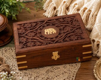 May include: A rectangular dark brown wooden box featuring an intricate floral carving on the lid and a central circular inlay depicting an elephant. The box includes a brass-colored metal latch and decorative brass-colored metal corner accents.