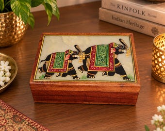 May include: A rectangular wooden box featuring a stone inlay on the lid. The inlay depicts two black elephants adorned with colorful red, green, and gold decorative blankets and harnesses, walking across a light-colored surface with green grass.