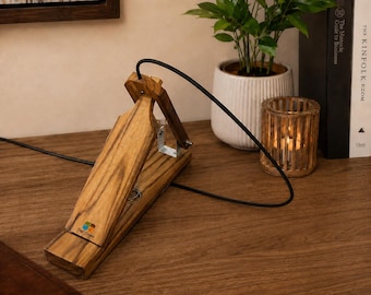 May include: A wooden foot pedal with a natural grain finish and a black power cord. The pedal features a small logo with blue, green, and orange squares. It is constructed with a hinged top section, a metal bracket, and a sturdy base for operation.