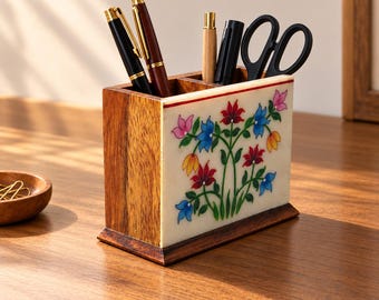 May include: A rectangular wooden desk organizer featuring a white stone front panel inlaid with a colorful floral pattern of red, blue, yellow, and pink flowers. The organizer has multiple compartments holding two pens, a wooden stylus, and scissors.