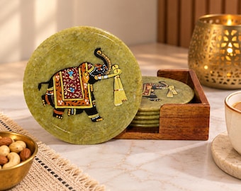 May include: A set of round, light green stone coasters featuring a painted black elephant with a red and gold decorative blanket. The coasters are stored in a square wooden holder, with one coaster displayed upright in front of the stack.