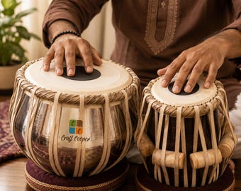 May include: A pair of tabla drums featuring a large metallic drum and a smaller wooden drum, both with white drumheads, black center patches, and beige tension straps. The metallic drum displays a logo with the text Craft Cooper.