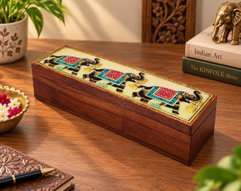 May include: A rectangular wooden box with a hinged lid featuring an inlaid panel. The panel displays three black elephants decorated with colorful red, blue, and gold patterned blankets, walking in a line against a light cream-colored background.