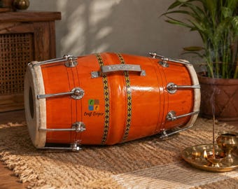 May include: A polished orange wooden dholak drum featuring silver-toned metal tuning hardware, a central carrying handle, and decorative black and yellow patterned bands. The drum has white skin heads and a small logo reading Craft Cooper.