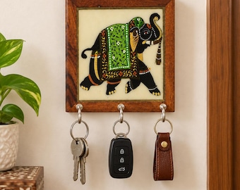 May include: A square wooden wall-mounted key holder featuring a central tile with a painted black elephant adorned in a green floral blanket. The frame is dark brown wood, and it includes three silver metal hooks holding two metal keys and a car fob.