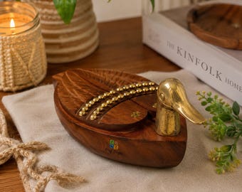 May include: A wooden duck-shaped box featuring a polished brass head and neck. The body is crafted from dark brown wood with carved wing details and two rows of small, round brass studs along the back. The brand name 'Emil Cooper' is printed.