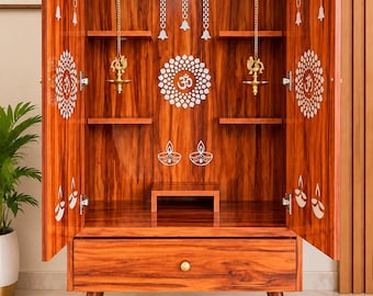 May include: A wooden home temple cabinet with two open doors, featuring a central Om symbol, hanging brass bells, and two brass oil lamps. The interior includes shelves and a small platform, while the exterior base has a single drawer and four legs.