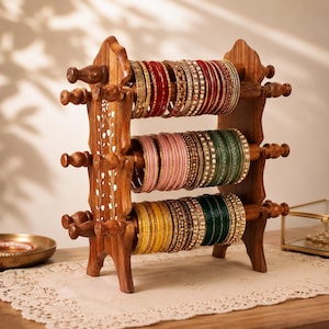 Puede incluir: Un soporte de madera para joyas de tres niveles con incrustaciones florales blancas en los paneles laterales. El soporte sostiene varias filas de brazaletes de varios colores, incluidos rojo, rosa, verde y amarillo, con detalles metálicos.