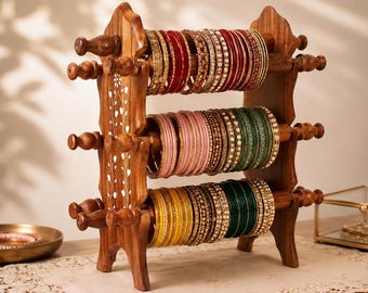 May include: A three-tiered wooden jewelry stand featuring intricate white floral inlay on the side panels. The stand holds multiple rows of bangles in various colors, including red, pink, green, and yellow, with metallic and beaded detailing.