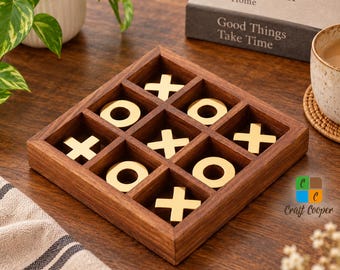 May include: A square wooden tic-tac-toe board with a dark brown finish, featuring nine grid compartments. The game includes five gold-colored metal X pieces and four gold-colored metal O pieces arranged within the slots of the wooden frame.