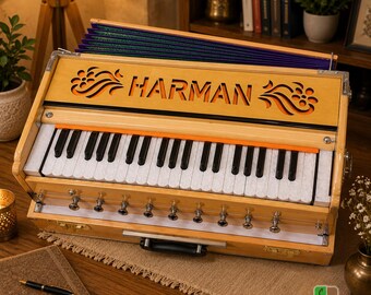 May include: A light-colored wooden harmonium musical instrument featuring a keyboard with white and black keys, an orange accent strip, and the word HARMAN laser-cut into the front panel. It includes a dark blue bellows and metal knobs.