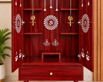 May include: A dark red wooden home temple cabinet with two open doors, featuring a central Om symbol, white floral patterns, and hanging brass bells. The interior includes shelves, two hanging brass oil lamps, and a bottom drawer with a gold knob.